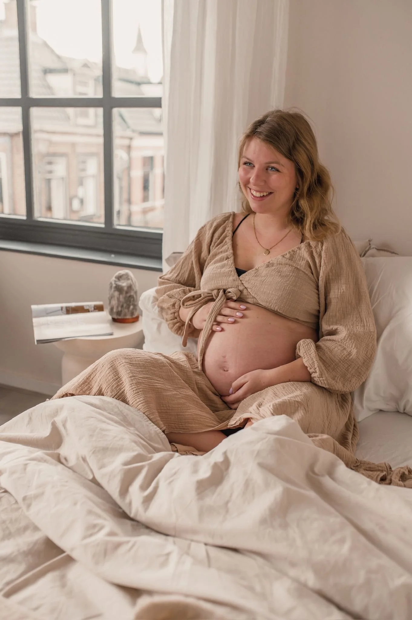 A pregnant woman sitting on a bed, smiling, wearing a tan dress and black undergarments, with her hand on her belly. There is a nightstand with a rock and an open book beside her, and a window with buildings outside in the background.