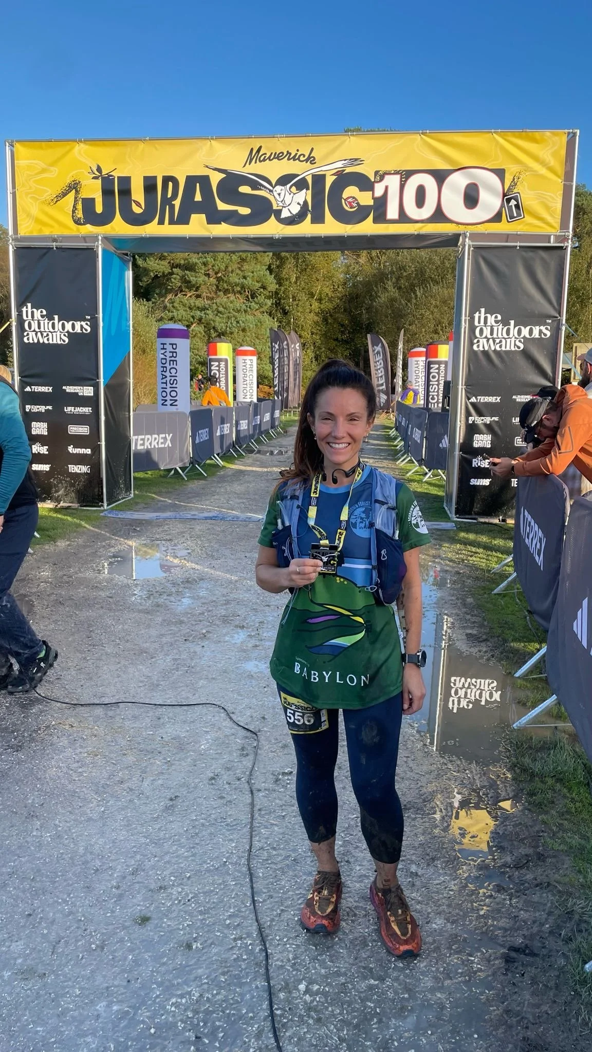 A woman smiling and holding a finisher medal at a trail race finish line for the Jurassic 100, with a yellow 'Jurassic 100' banner overhead and muddy trail behind her.