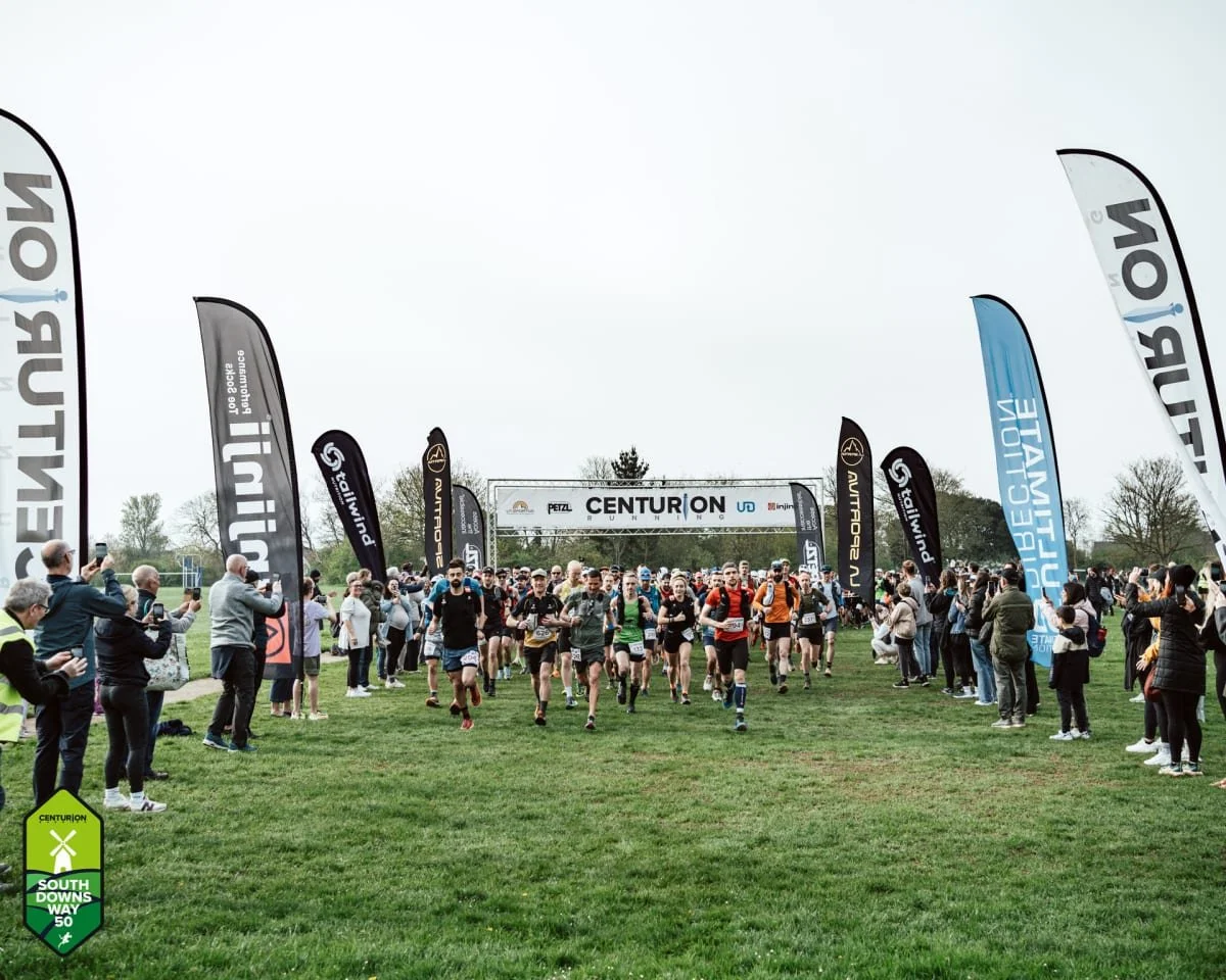 Runners participating in a marathon at the starting line outdoors, with spectators and photographers on sides, and a banner overhead reading "Centurion Running".