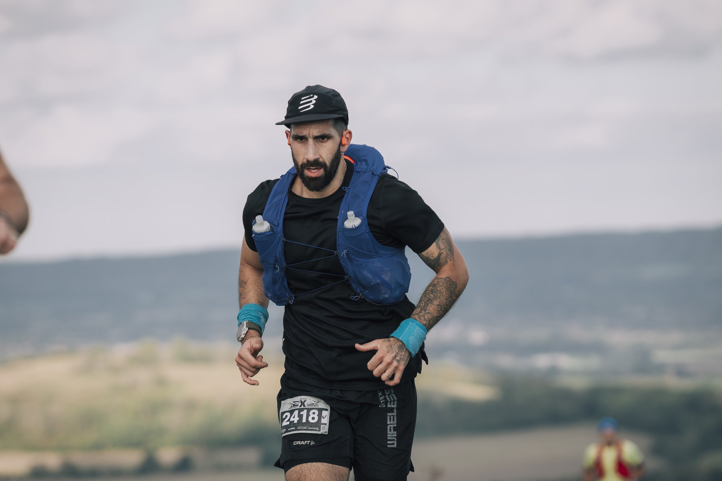 Male marathon runner with tattooed arms, wearing a black shirt, shorts, a blue hydration vest, and a black cap, running outdoors with a cloudy sky and landscape in the background.