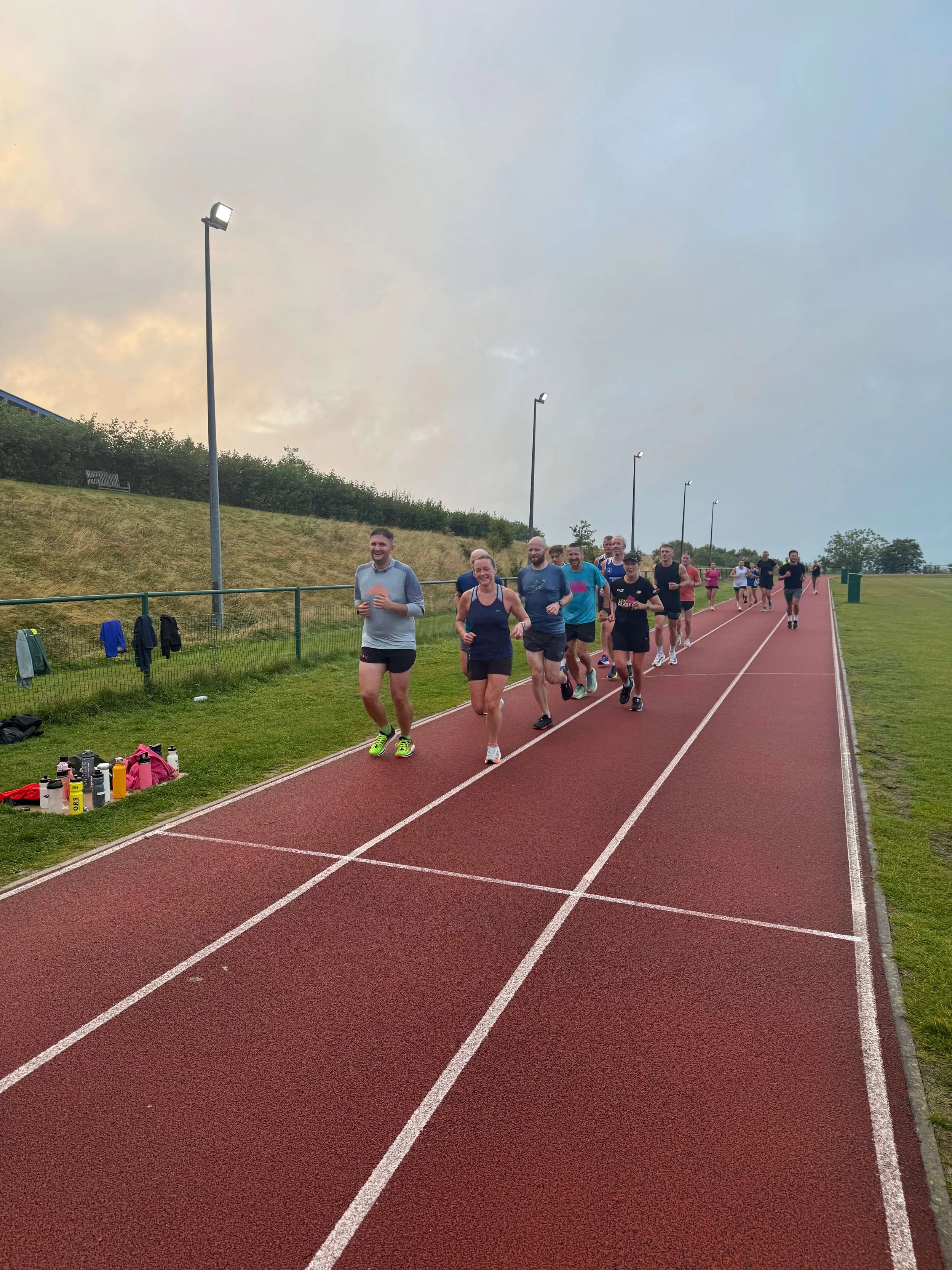 A group of people jogging on a red outdoor running track with lampposts and grassy areas in the background.