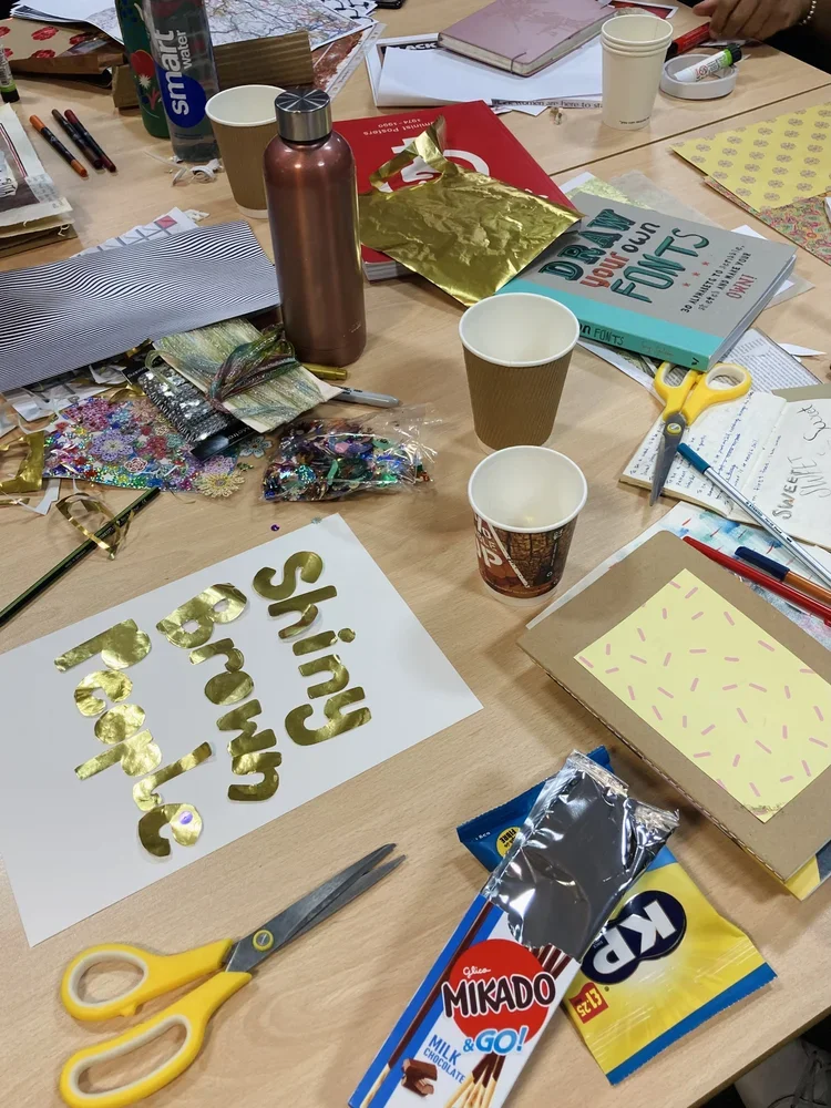 Table with crafting supplies including scissors, a gold 'Happy Birthday' sign, chocolates, paper cups, notebooks, pens, scissors, and wrapping paper.