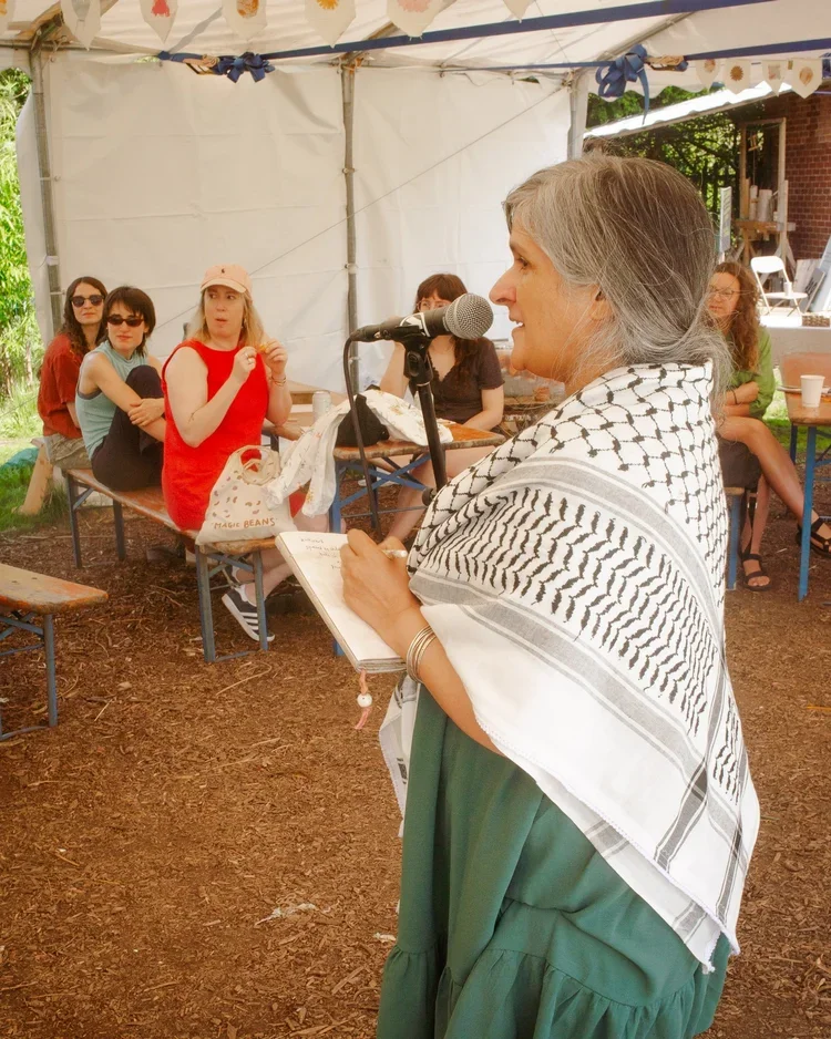 A woman with gray hair dressed in green, holding a notebook, speaking into a microphone at an outdoor gathering under a tent, with several women sitting at a table listening.
