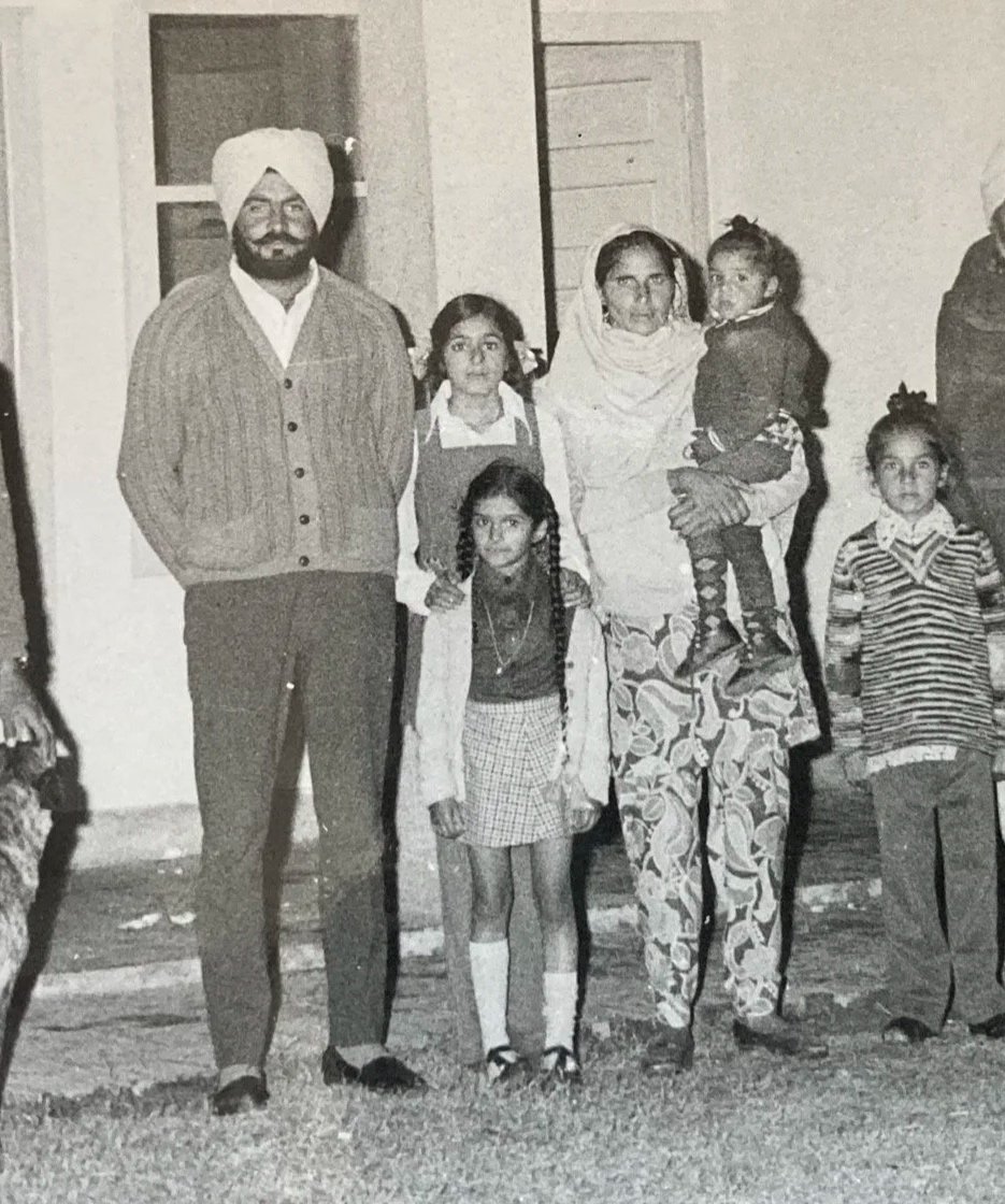 A black and white photo of a Sikh family group standing outdoors, including a man wearing a turban, a woman with a scarf, and five children in casual and traditional clothing.