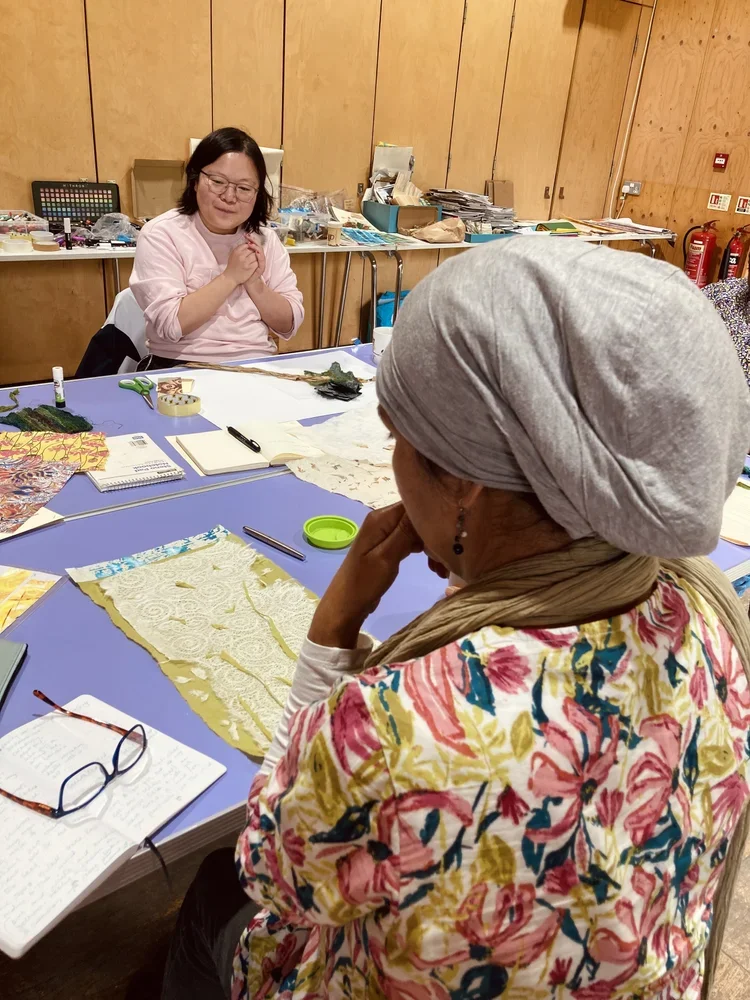 Two women sitting at a table with colorful fabric, notebooks, and sewing supplies, engaged in a craft or sewing activity in a wood-paneled room.