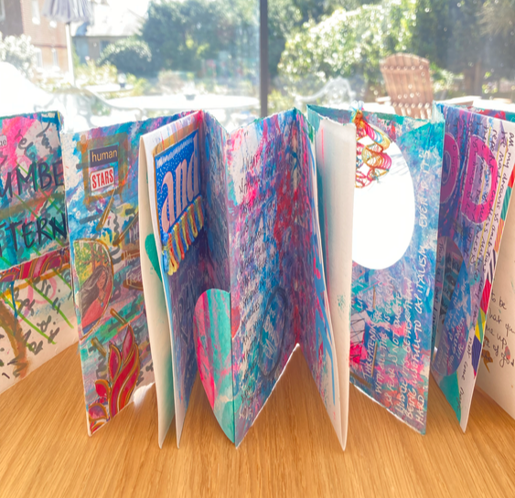Open colorful scrapbook with various decorated pages, placed on a wooden surface with sunlight coming through a window in the background.