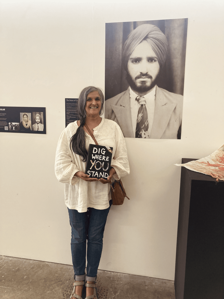Woman with long gray hair holding a sign that says 'Dig Where You Stand' in front of a large black-and-white portrait of a man in a turban, suit, and tie at an art exhibit or gallery.