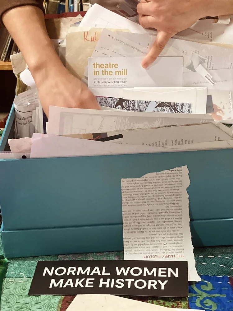 A stack of papers and booklets on a table, with a person pointing at a brochure titled 'theatre in the mill'. In the foreground, there is a sign that reads 'NORMAL WOMEN MAKE HISTORY'.