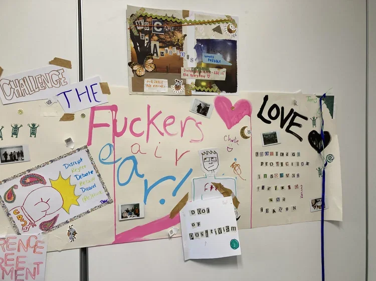 A whiteboard decorated with various colorful posters, drawings, and handwritten messages. Prominent words include "The Fuckers" in pink, "LOVE" with a black heart, and displays related to love, positivity, and challenges. There are small photos, dood