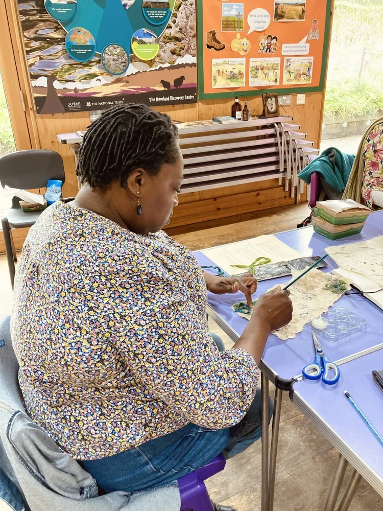 A woman with short, dreadlocked hair working on an arts and crafts project at a table in a room with wooden walls. She is using a brush on a piece of fabric, and materials like scissors, paper, and colorful textiles are on the table. In the backgroun