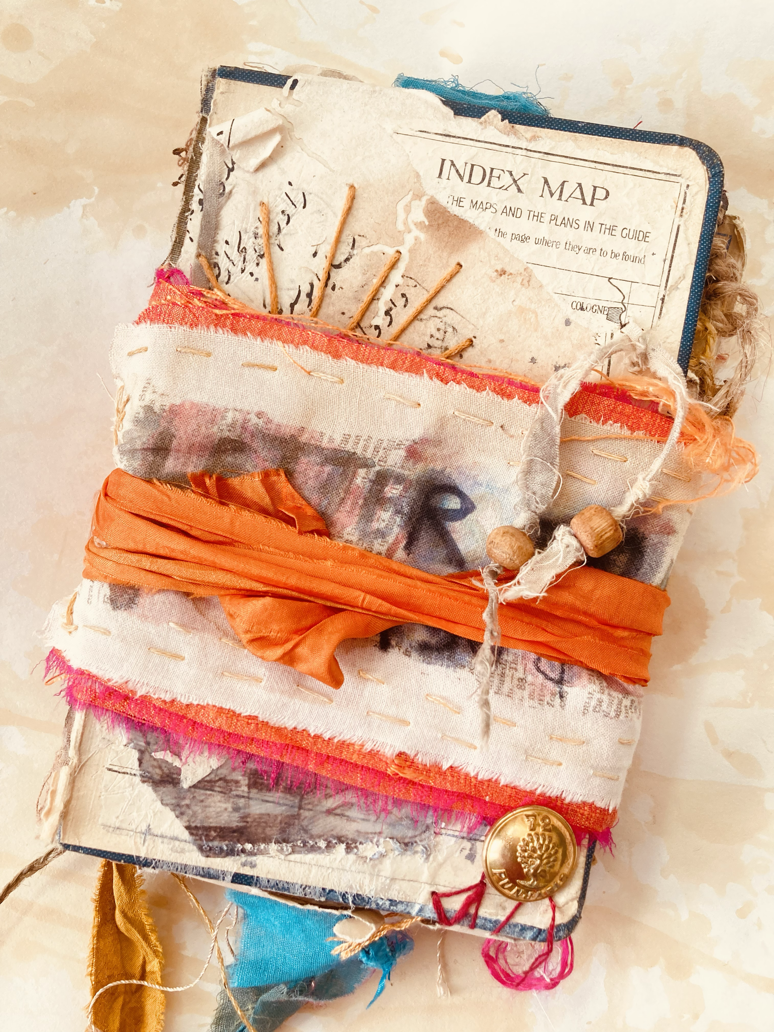 A worn, fabric-covered book with tattered pages and various fabric scraps, including an orange ribbon, attached threads, and a decorative button.