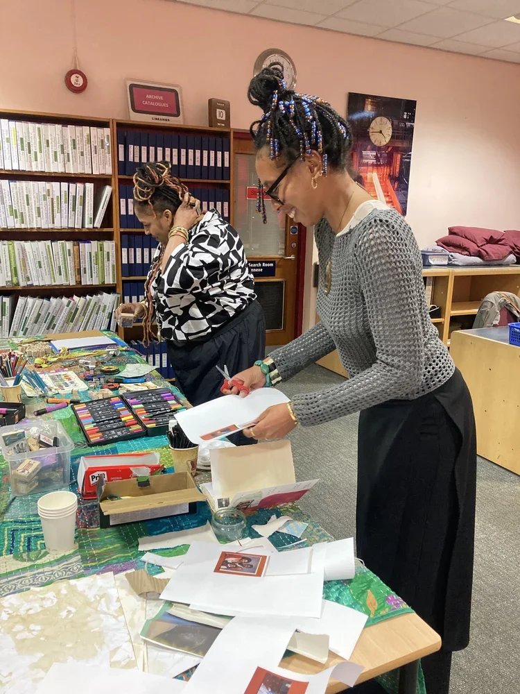 Two women with colorful braided hair and glasses working at a craft table in a library or office, surrounded by various craft supplies, papers, and drawings.