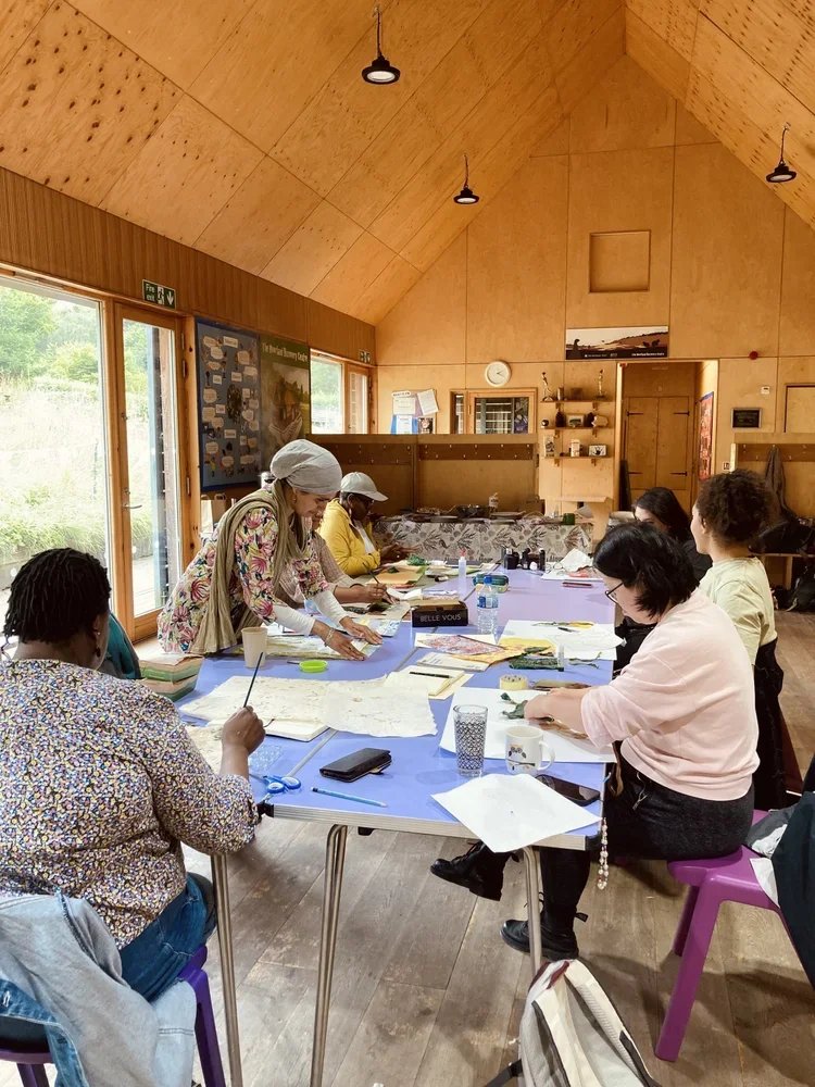 A group of women gathered around a table in a wooden community room or classroom, working on maps, books, and crafts with windows showing greenery outside.