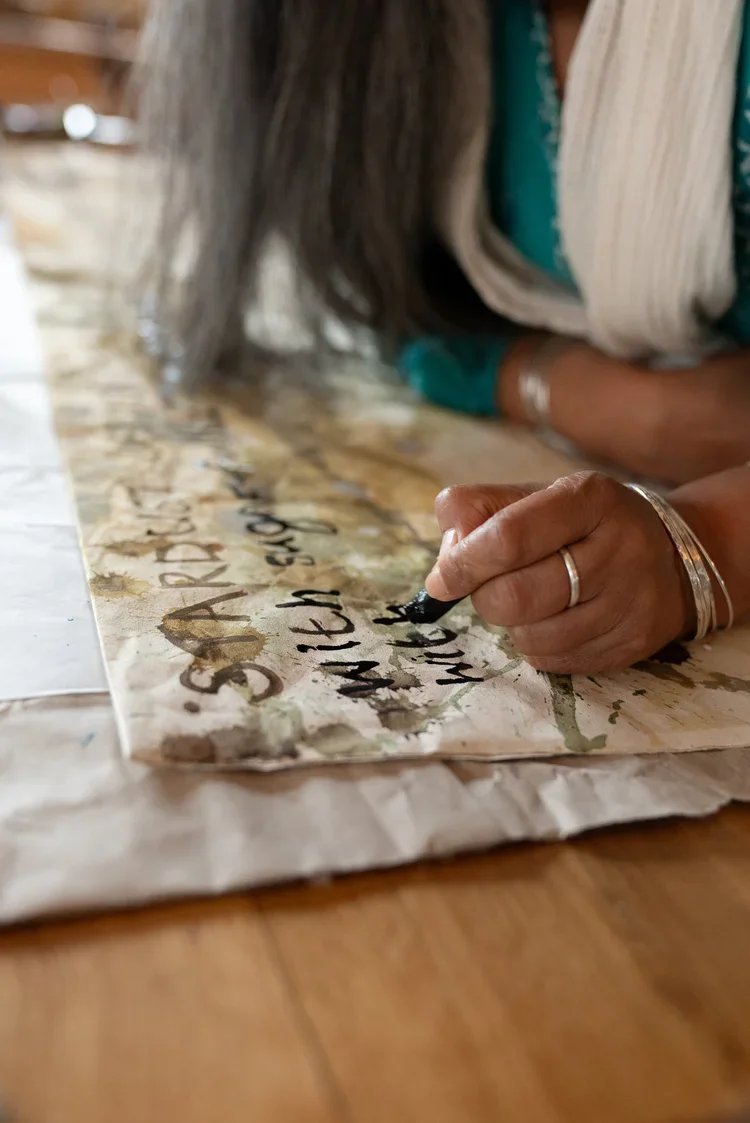 A person writing on a piece of paper with a black marker.  The person's hand is adorned with jewelry and they are wearing a teal long-sleeve shirt and a scarf.