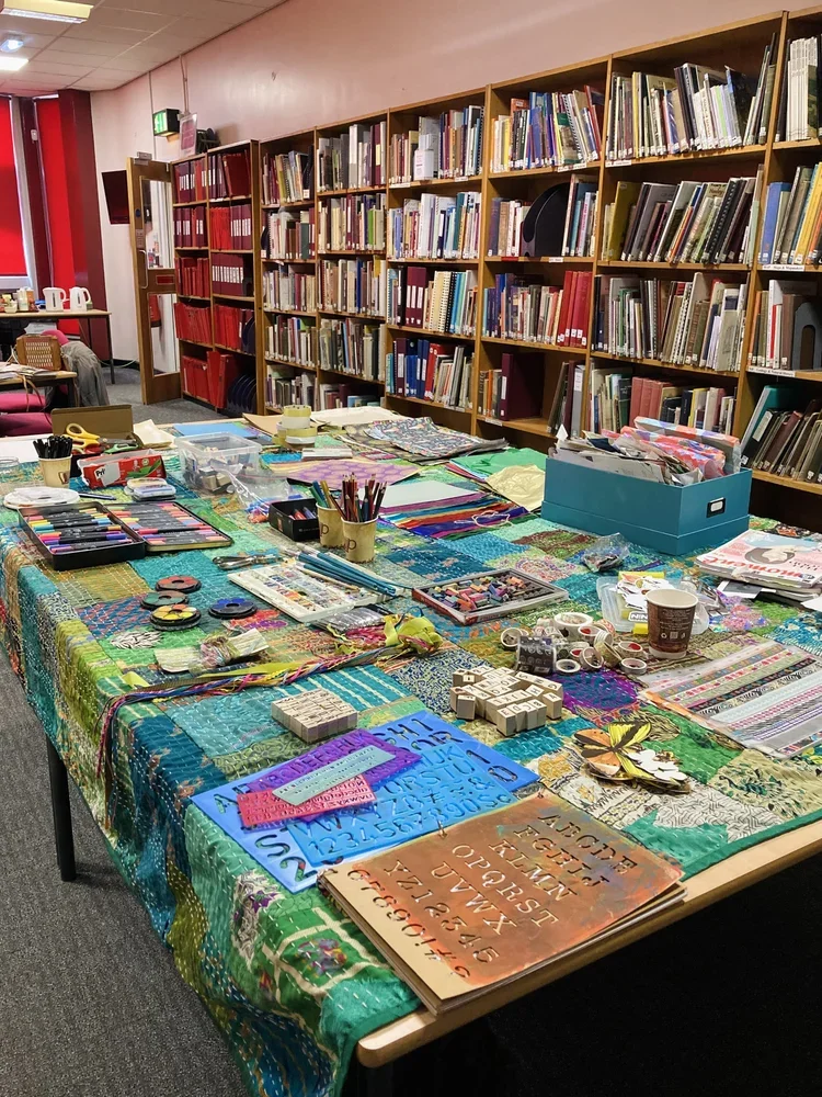 A table covered with colorful quilting supplies, including fabric, rulers, scissors, and tools, set in front of wooden bookshelves filled with books in a library or craft room.