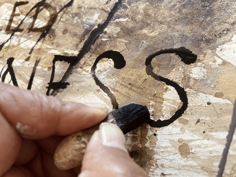 A person is using a black crayon with a wooden handle to trace and fill in the letter 'S' on a weathered, brown surface, likely a piece of wood or paper.