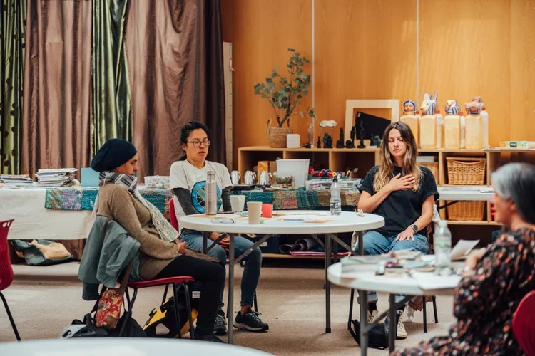 Group of women sitting in a circle during a meditation or mindfulness session in a cozy room with wooden walls, shelves with decorations, and a table with books and personal items.