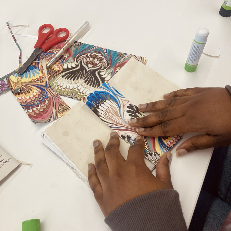 Person folding marbled paper on a white table with scissors, glue sticks, and a colorful paper sample.