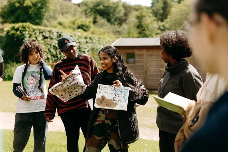 Group of five young people outdoors, one holding a sign with handwritten message, others holding notebooks, smiling, with a wooden building and trees in the background.