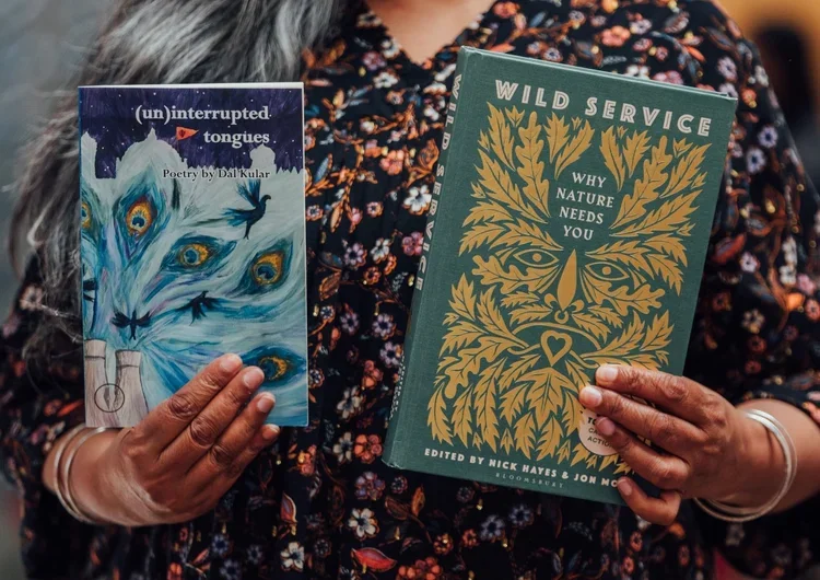 Person holding two books, one titled '(un)interrupted tongues' with an abstract blue and white cover featuring peacock-like feathers, and the other titled 'Wild Service' with a green cover and a stylized leaf and face design.