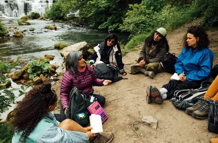 Group of six women sitting on a dirt path by a river, engaged in a conversation, surrounded by lush greenery.
