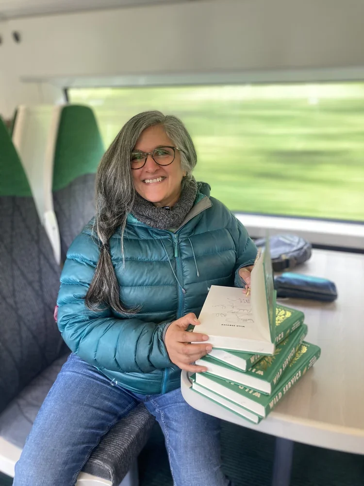 A smiling woman with gray hair and glasses, wearing a teal puffer jacket, sitting on a train seat, holding a stack of books and opening one.