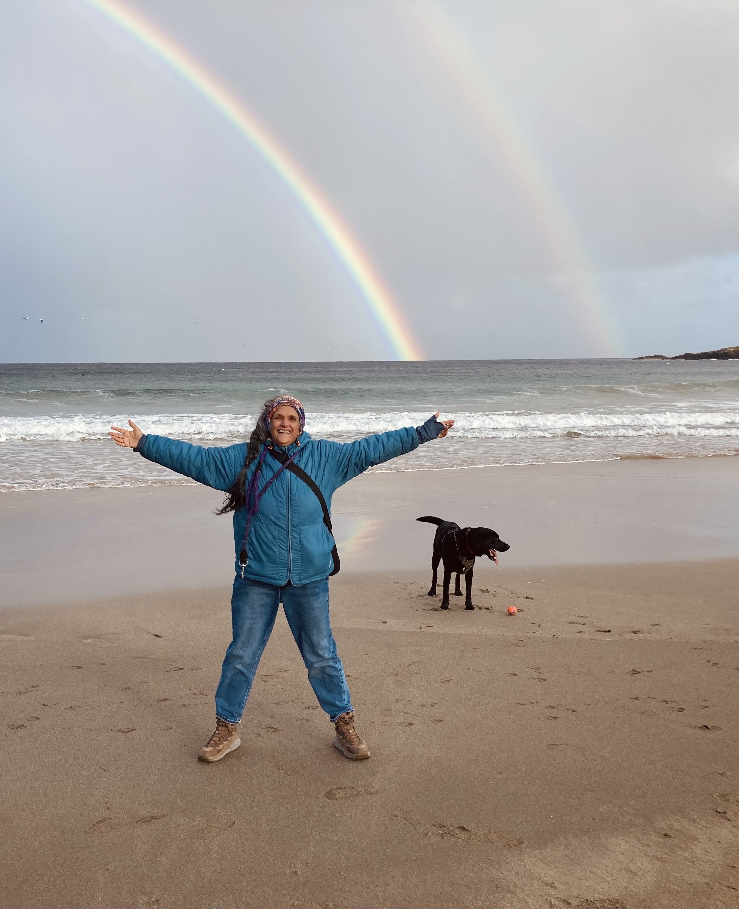Brown woman in a blue jacket and jeans on the beach with arms wide open, smiling, with a black dog nearby and a double rainbow in the cloudy sky over the ocean.
