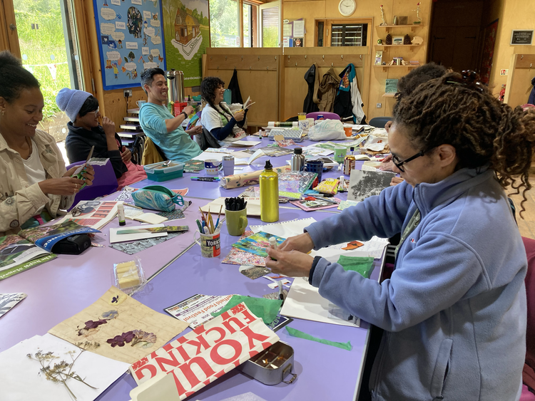 A group of people sitting around a purple table in a classroom, engaging in arts and crafts. There are various supplies like paper, scissors, glue, and markers on the table, with some participants smiling and chatting.