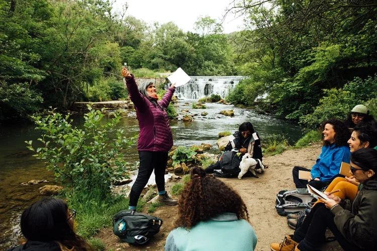 Group of people seated by a river, listening to a woman in a purple jacket holding a paper, engaging with the group during a nature outdoor activity.