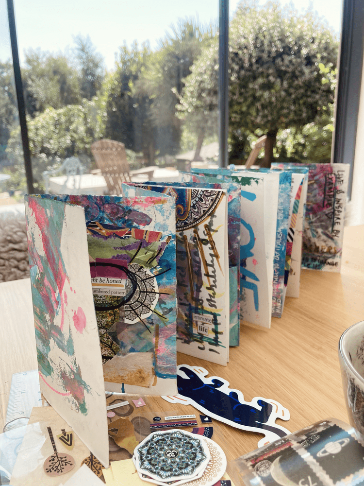 A colorful, artistic paper collage or booklet displayed on a wooden table near a glass window with a sunny outdoor garden scene.