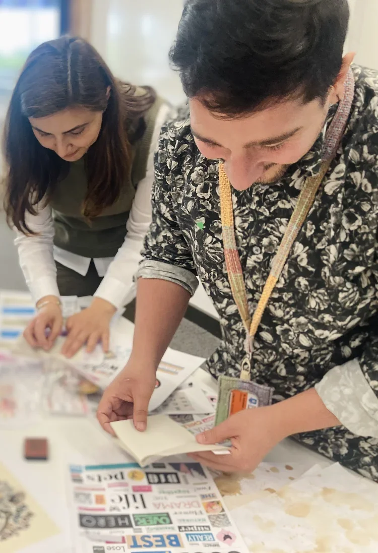 Two people looking at newspapers at a table, with various printed materials spread out, engaged in reading or comparing papers.