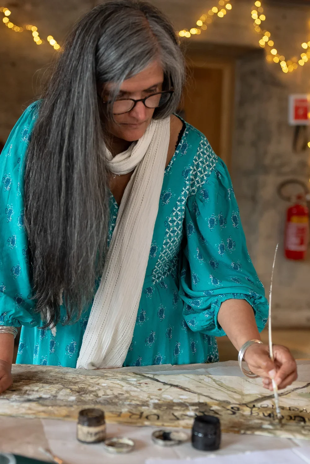 Woman with long gray hair and glasses wearing a teal patterned dress and a white scarf, working on a large watercolor painting at a table, with a string dipped in black ink or paint. Decorative string lights and a fire extinguisher are visible in the