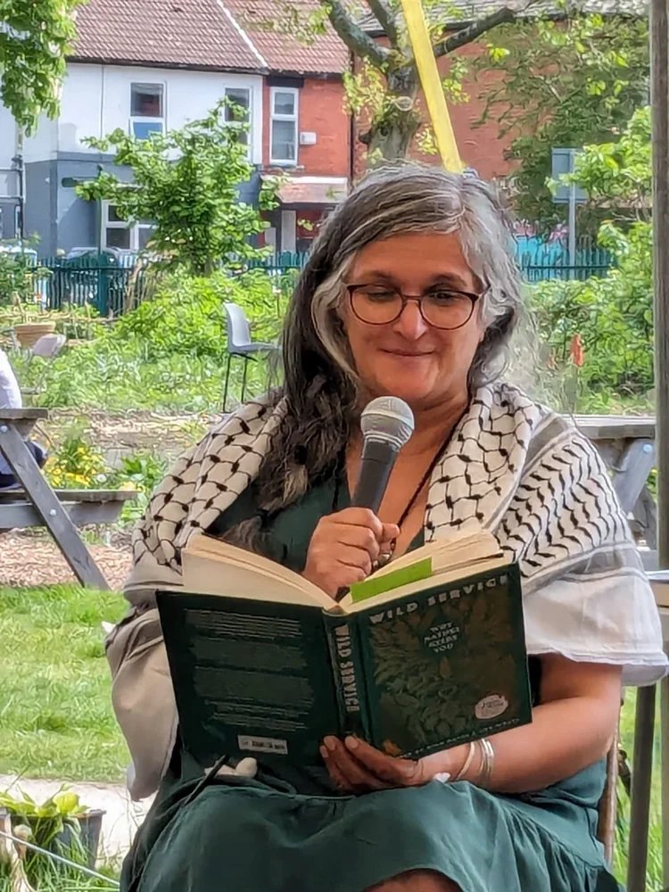 A woman with wavy gray hair and glasses sitting outdoors, reading a book titled 'Wild Service' while holding a microphone.