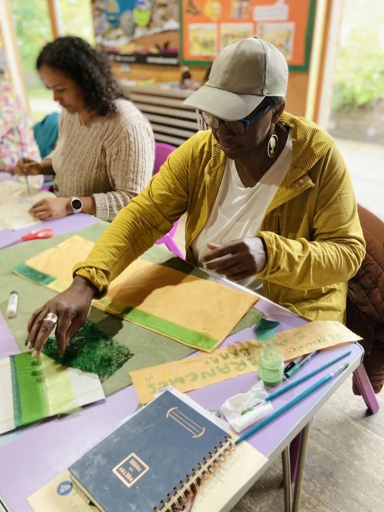 Two women working on art projects at a table in a classroom, with various art supplies and colorful posters on the wall.