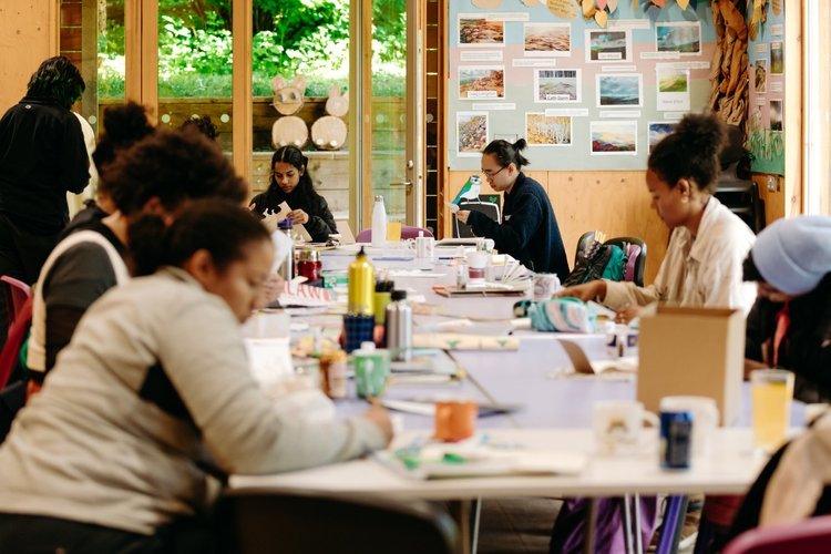 People sitting around a large table engaged in arts and crafts activities inside a room with large windows and nature outside. There are art supplies on the table and pictures on a bulletin board in the background.