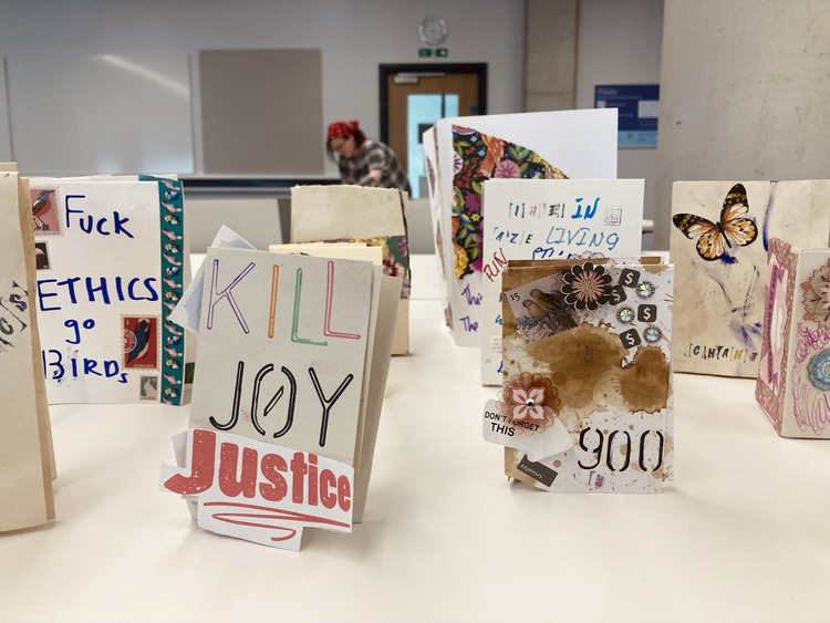 Collection of handmade zines with various messages and artwork displayed on a table, with a blurred person in the background.
