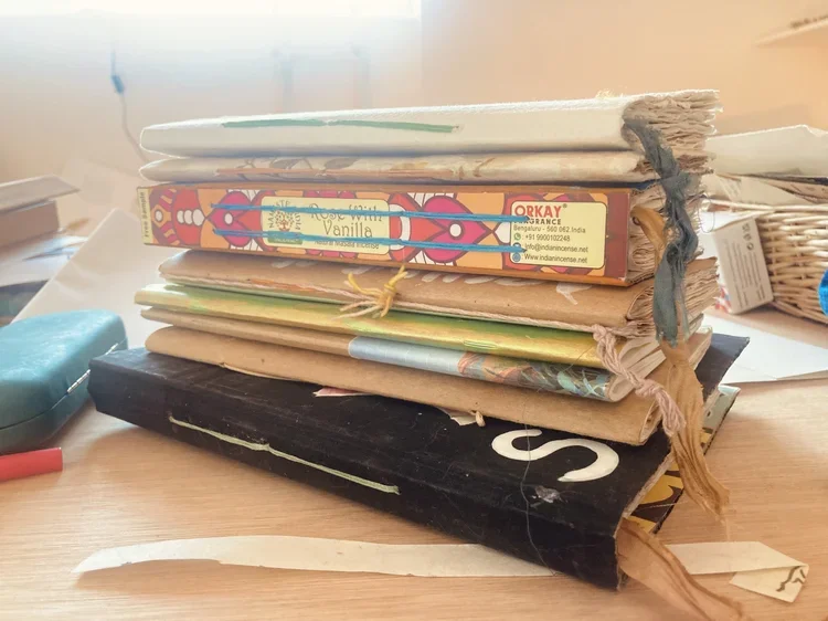 Stack of old, worn books and notebooks on a wooden desk, with loose papers and other items nearby.