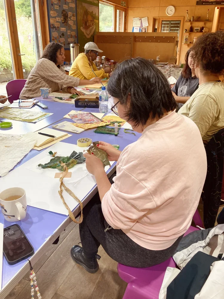 A group of women sit around a table engaged in arts and crafts activities. The table has various craft supplies, water bottles, and a mug. The room has wooden walls and large windows letting in natural light.