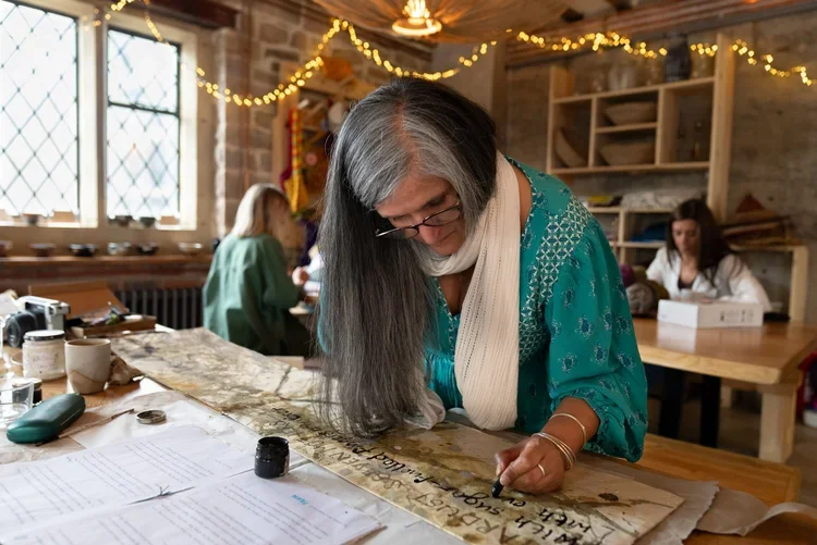 A woman with long gray and black hair, wearing glasses, a teal blouse, and a cream scarf, is working on an old document or scroll at a wooden table decorated with books, jars, and writing tools. In the background, people are working in a cozy room de