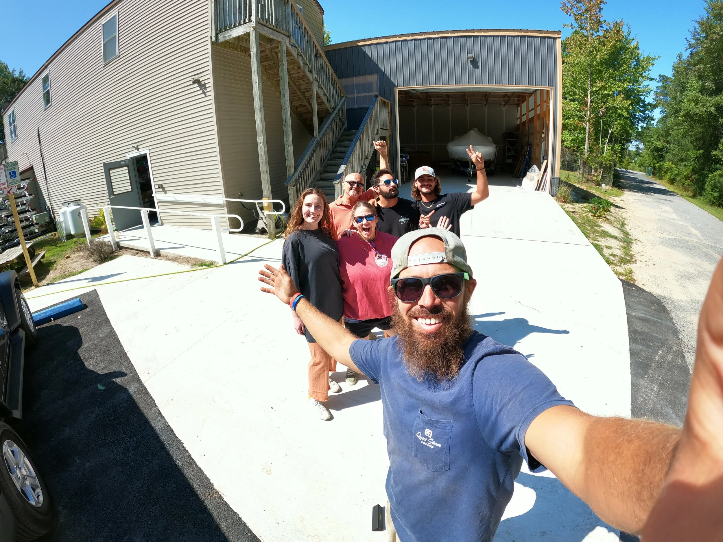 A group of six people smiling and taking a selfie outside near a building with a blue sky, green trees, and a boat stored in a garage in the background.