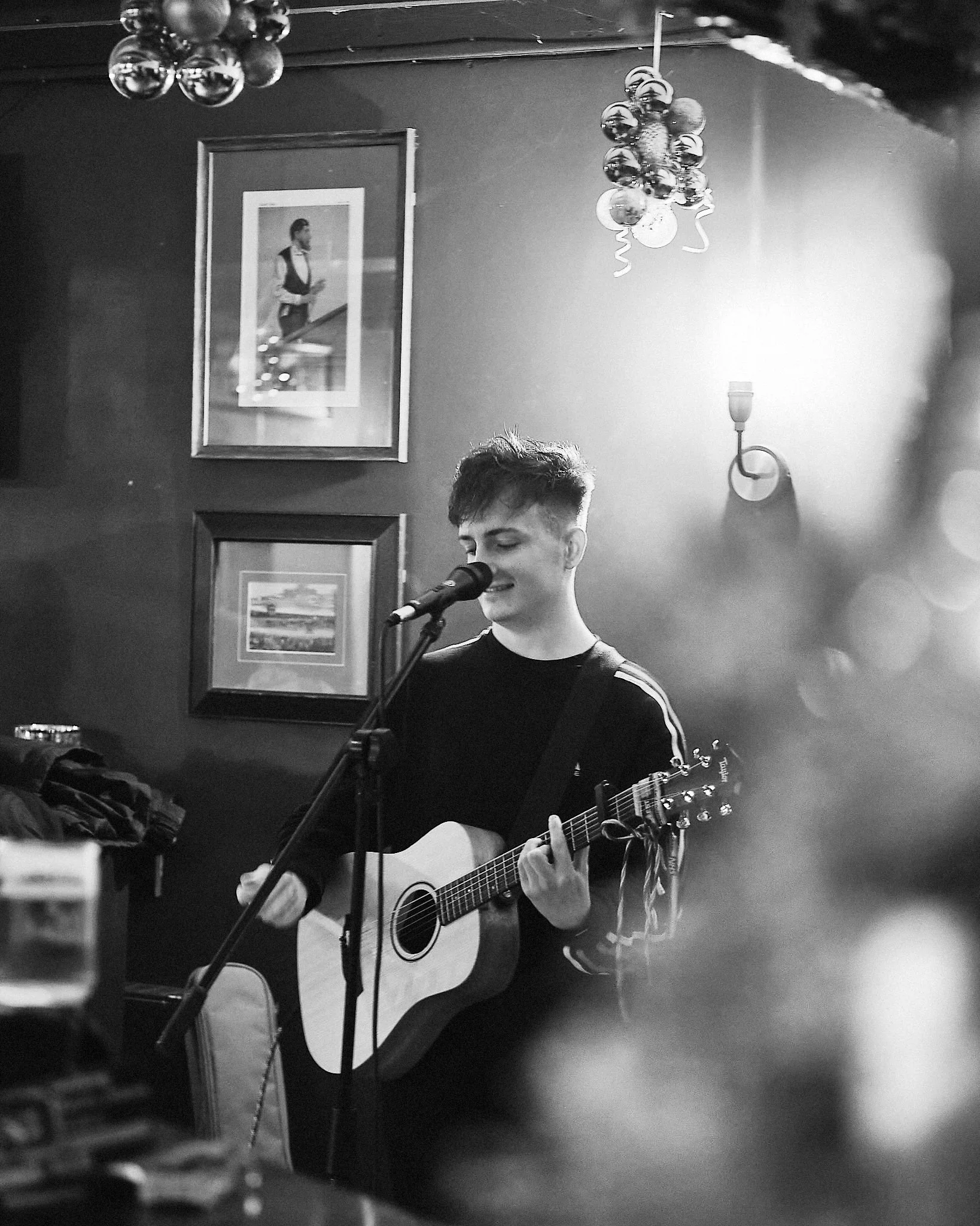 A young man with short, styled hair playing an acoustic guitar and singing into a microphone in a dimly lit bar or cafe with framed pictures on the wall and a chandelier hanging from the ceiling.