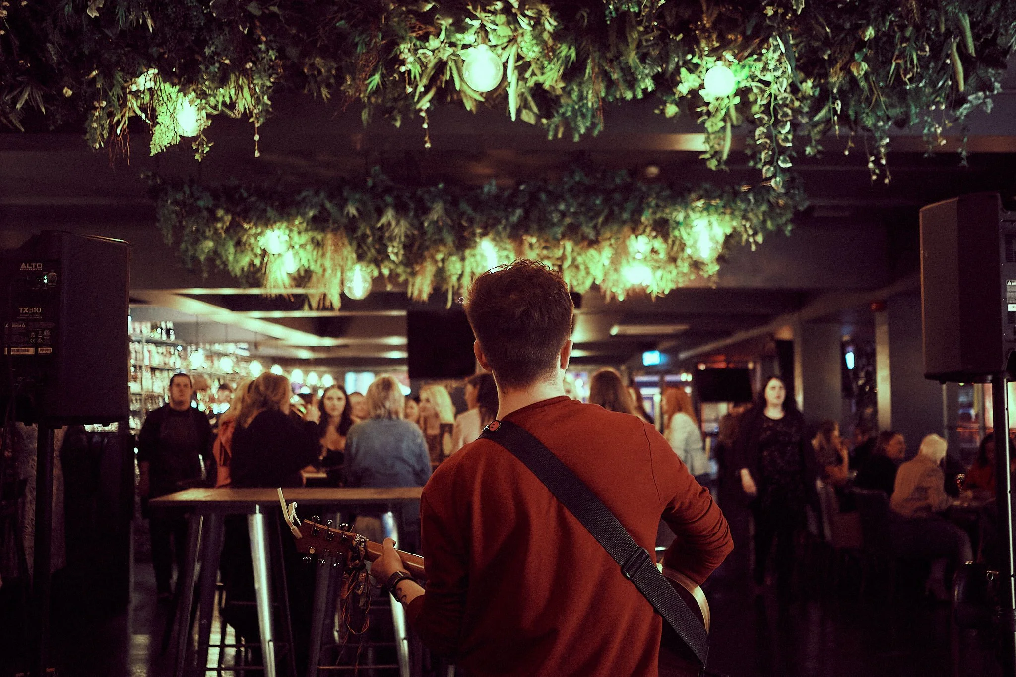 A man with a guitar performing on stage in a dimly lit bar or restaurant, with an audience watching. The ceiling is decorated with hanging greenery and lights.