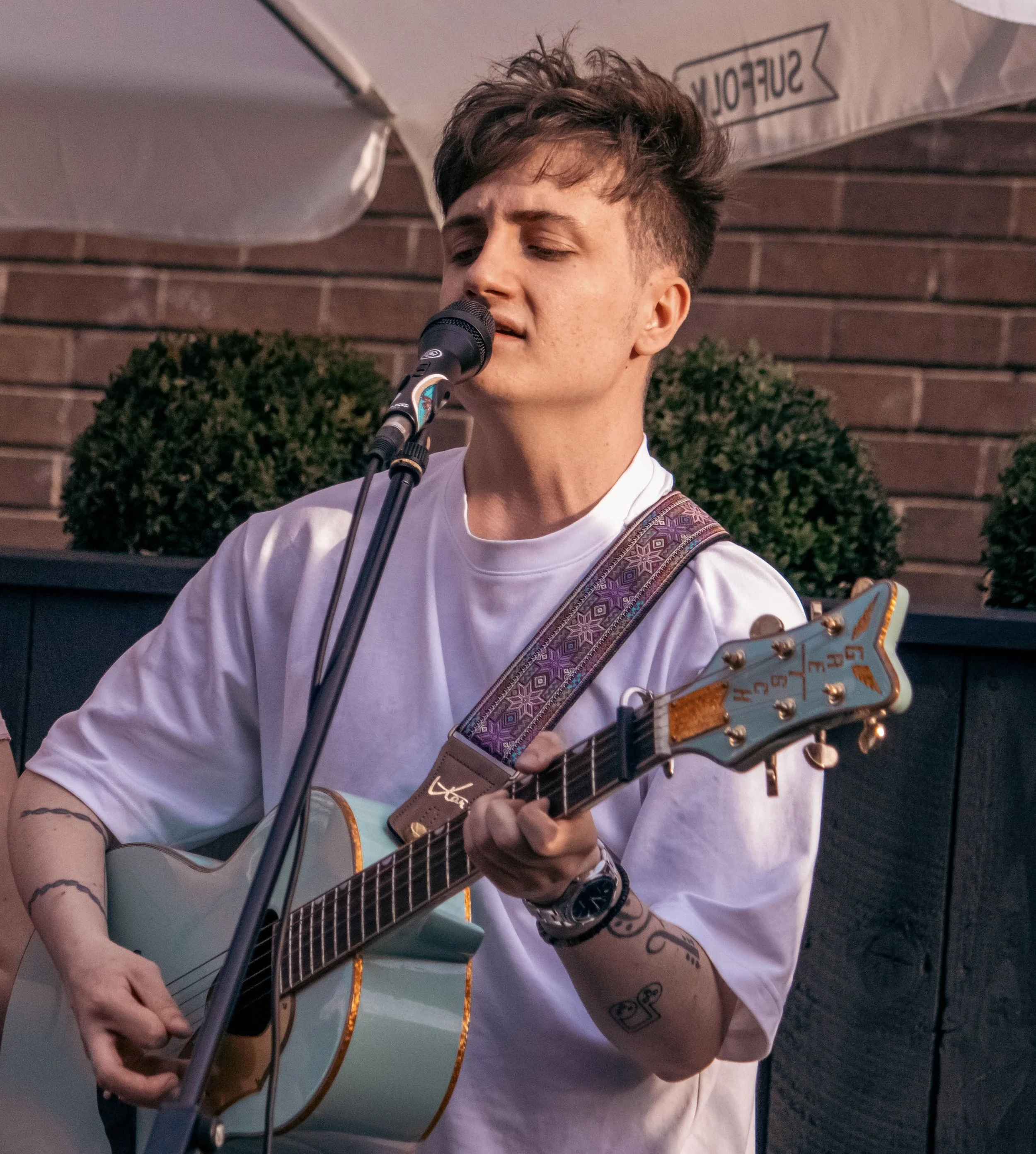 Young man singing into a microphone while playing an acoustic guitar outdoors, with a brick wall and greenery in the background.