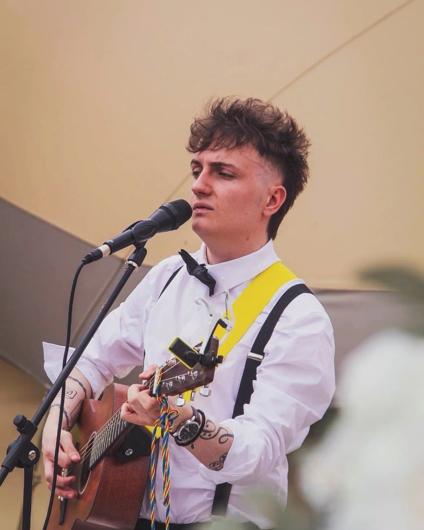 A young man with curly hair wearing a white shirt and black bow tie, playing an acoustic guitar and singing into a microphone, with tattoos on his arms.