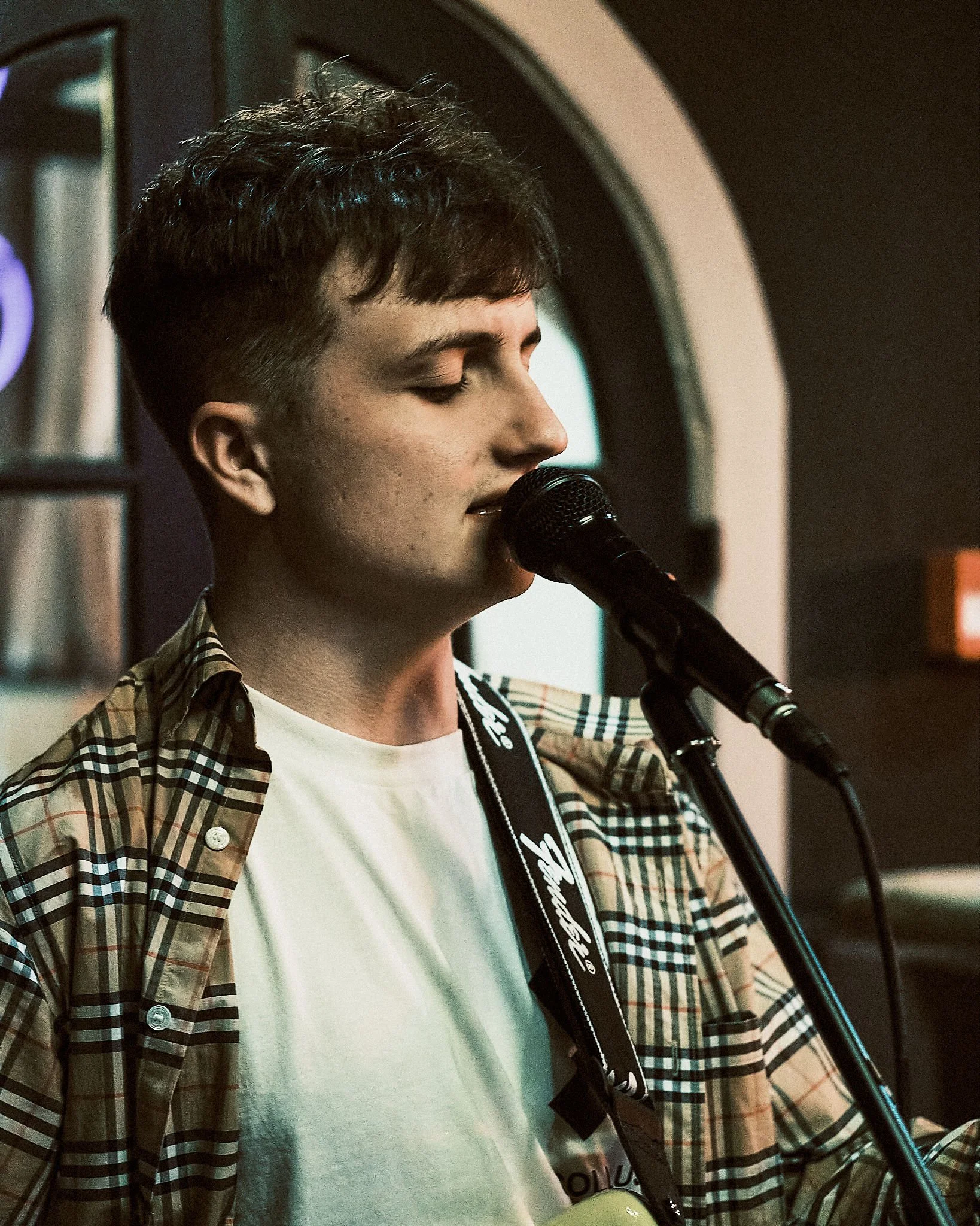 A young man with dark hair singing into a microphone while playing guitar in a dimly lit room.