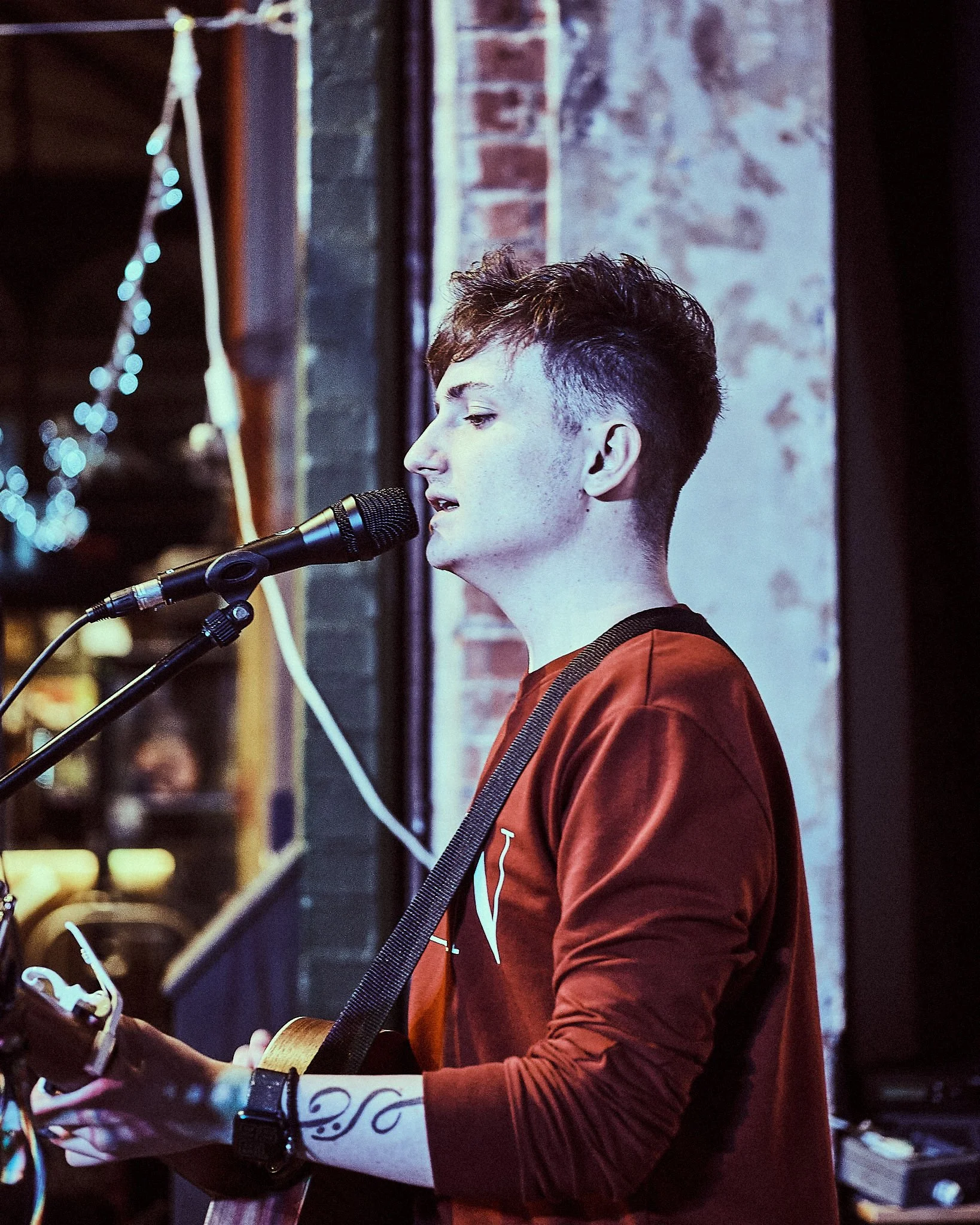 A young man with short hair singing into a microphone while playing an acoustic guitar, standing in front of a brick wall in a dimly lit indoor setting.