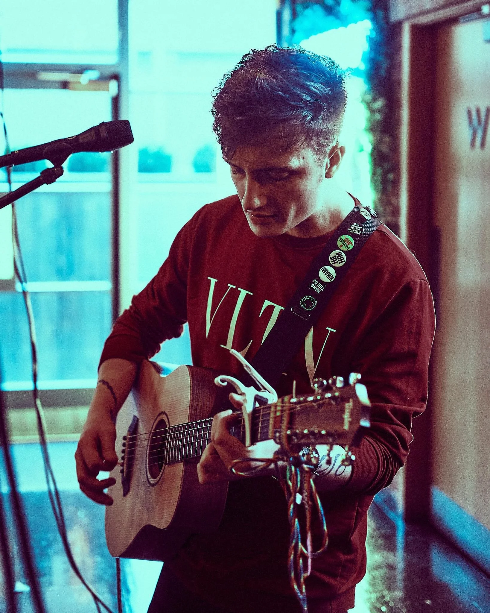 A young man playing an acoustic guitar near a microphone, wearing a red sweatshirt, indoors with blue lighting.