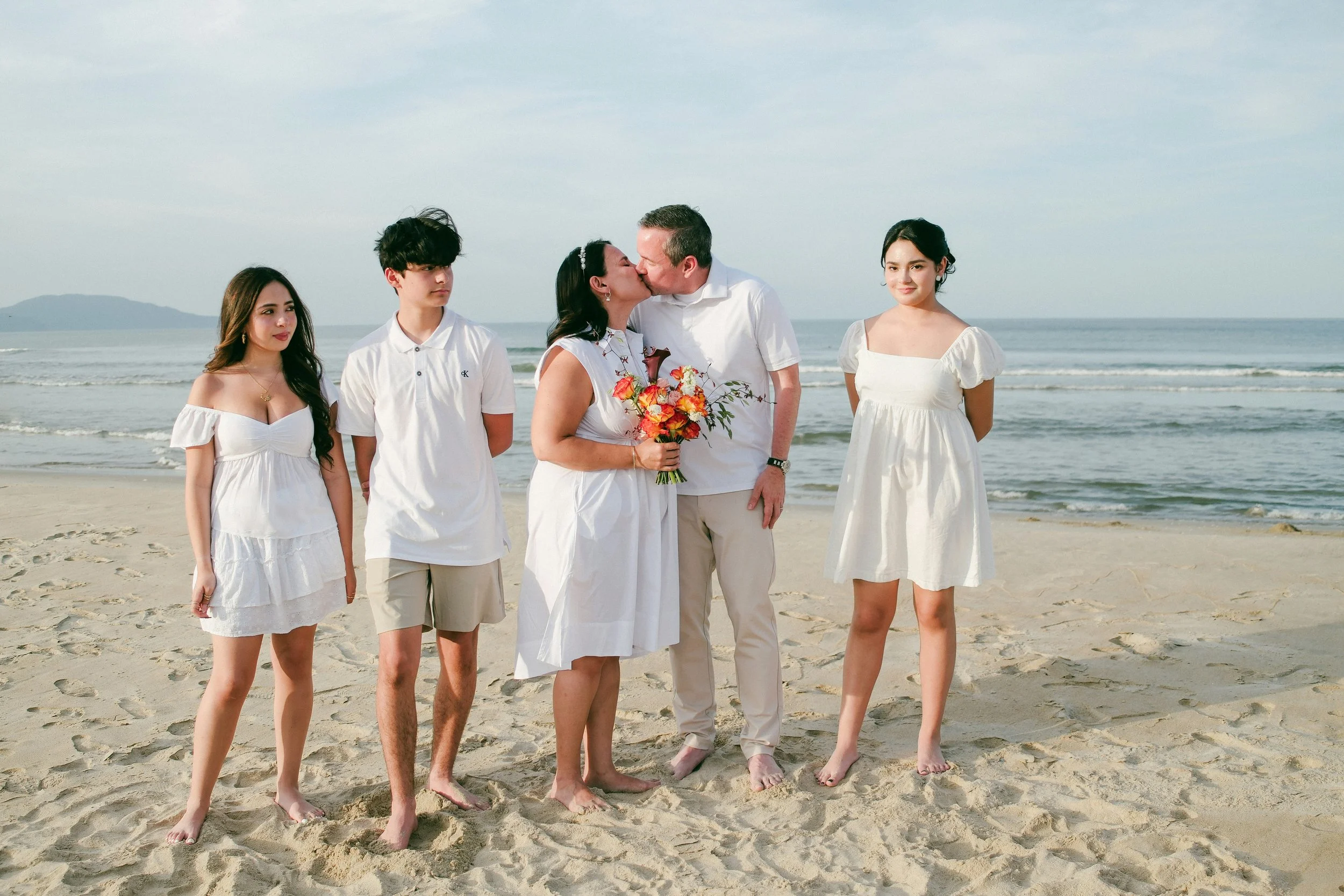 Couple sharing a kiss during a beach vow renewal ceremony, standing with their children by the sea.