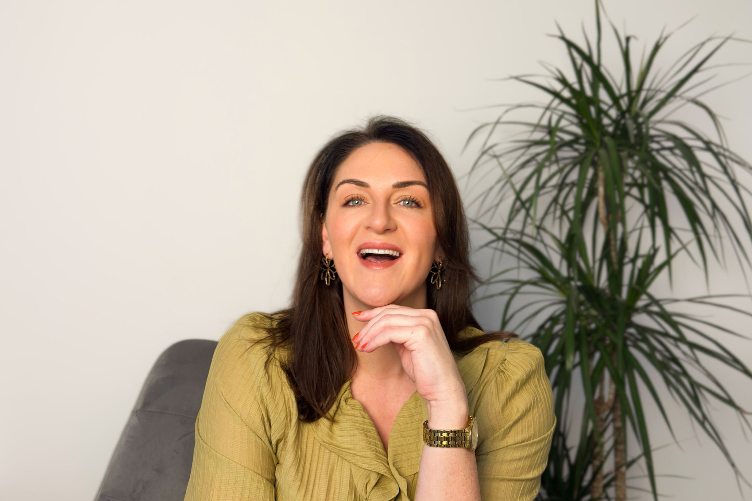 Sarah Precious, wedding celebrant at Sunday Best Ceremonies, seated indoors and smiling warmly, with a leafy plant in the background.