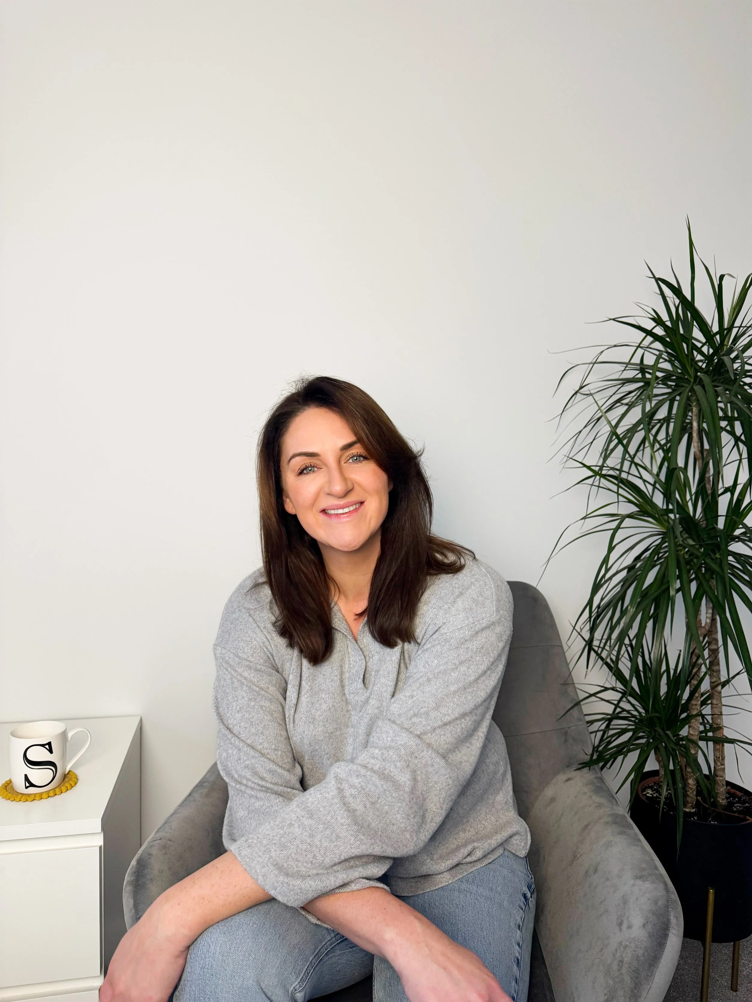 Meet Sarah Precious celebrant and founder of Sunday Best Ceremonies who has brown hair and blue eyes sitting on a grey chair, smiling at the camera, wearing a light grey sweater and jeans, with a tall green plant behind her and a white side table wit